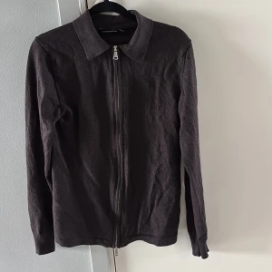 J lindberberg tröja - Snygg mörkbrun kofta från J.Lindeberg med hel dragkedja framtill och klassisk krage. Långärmad modell i mjukt stickat material, perfekt för lager-på-lager-stil. Passar dig som gillar stilrena och enkla plagg med en twist.