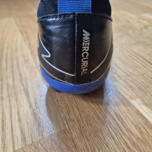 Nike Mercurial Air Zoom fotbollsskor svart/blå - Säljer ett par Nike Mercurial Air Zoom fotbollsskor i svart med blå detaljer och vit Nike-logga. Skorna har en strumpliknande passform, kardborreband över vristen och blå yttersula. Perfekta för dig som vill ha grymt grepp och stil på planen.