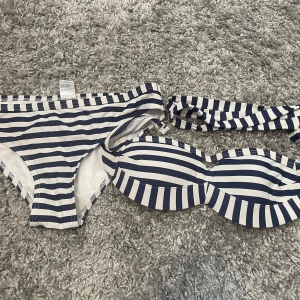 Randig bikini från Stadium, stl 36 - Snygg bikini med marinblå och vita ränder från Stadium. Överdelen är bandeau-modell med avtagbara axelband och bygel, underdelen är klassisk trosa. Perfekt för stranden eller poolen. Materialet är mjukt och stretchigt syntet.
