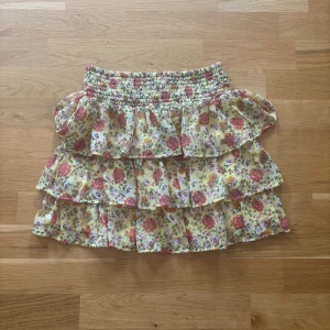 Blommig volangkjol från Lindex - Supersöt kort volangkjol från Lindex med blommigt mönster i rosa, gul och grönt på ljusgul botten. Kjolen är i storleken 134/140 men passar dig som annars har xs eller s.