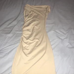 Beige kort klänning från H&M - Snygg beige kortklänning från Divided med draperad effekt på ena sidan. Klänningen är ärmlös och har en figurnära passform som framhäver siluetten. Perfekt för dig som gillar stilrena och trendiga plagg.
