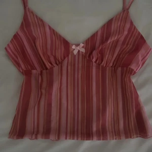 Randig rosa linnetopp med rosett - Supersöt randig linnetopp i olika rosa nyanser med tunna axelband och en liten rosett framtill. Toppens tyg är lätt och luftig, perfekt för varma dagar. Den har en feminin v-ringning och smickrande passform.