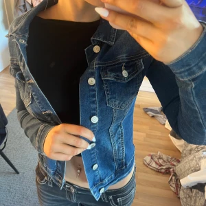 Mörkblå jeansjacka  - Snygg mörkblå jeansjacka från Sincerely Loved i klassisk modell med vita knappar och två bröstfickor med lock. Jackan har långa ärmar och kontrastsömmar som ger en cool look. Perfekt att slänga över en t-shirt eller hoodie.
