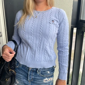 Ljusblå kabelstickad tröja Bondelid - Supersnygg ljusblå kabelstickad tröja från Bondelid med rund hals och broderad logga på bröstet. Tröjan har lång ärm och ribbade muddar för en klassisk look. Perfekt att styla med jeans eller kjol för en chill vibe.