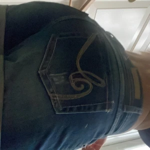 Blå jeansshorts med broderad ficka - Snygga blå jeansshorts med uppvikta ben och coolt broderi i beige på bakfickorna. Klassisk femficksmodell med bälteshällor och slitna detaljer för en avslappnad vibe. Perfekta för varma dagar när du vill ha en chill men ändå stilren look.