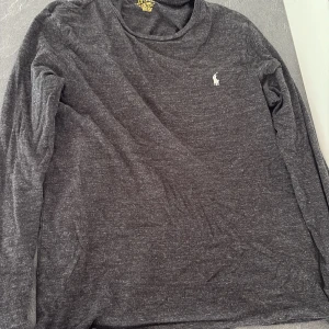 Mörkgrå långärmad tröja från Polo Ralph Lauren - Mörkgrå långärmad tröja från Polo Ralph Lauren med klassisk vit broderad logga på bröstet. Tröjan har en enkel, stilren design och är tillverkad i mjuk bomull. Perfekt för dig som gillar en clean och avslappnad look.