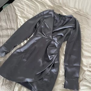 Mörkgrå satin klänning från Zara - Snygg mörkgrå kortklänning i satin med omlott-look och långa ärmar. Klänningen har markerad midja, breda manschetter med knappar och en elegant drapering framtill. Perfekt för dig som gillar stilrena och trendiga plagg med lyxig känsla.