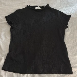 Svart blus med volangkant Kappahl XL - Svart blus från Kappahl i storlek XL. Toppen har korta ärmar med volangkant och en lätt rynkad struktur i tyget. Hög hals med vågig kant ger en söt touch. Perfekt att styla med jeans eller kjol för en trendig look.