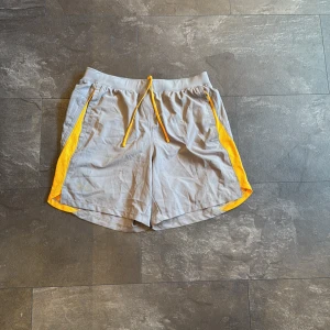 Grå sportshorts med gula detaljer - Snygga grå sportshorts från Under Armour med gula meshpaneler längs sidorna och matchande gul snörning i midjan. Shortsen har en avslappnad passform och är perfekta för träning eller aktiva dagar. Lätta och bekväma med elastisk midja och praktiska fickor.