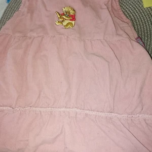 Rosa klänning Disney Baby med Nalle Puh - Supersöt rosa kortklänning från Disney Baby i mjuk manchester. Klänningen har ett broderat motiv av Nalle Puh på bröstet och knappar på sidan för enkel påklädning. Perfekt för små barn som gillar klassiska figurer.