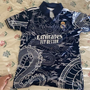 Real Madrid blå draktröja Adidas - Snygg Real Madrid fotbollströja från Adidas i mörkblått med ett stort drakmönster i silver. Tröjan har korta ärmar, klassisk krage och klubbmärke på bröstet. Materialet är lätt och andas, perfekt för match eller träning. Emirates Fly Better-logga på framsidan.