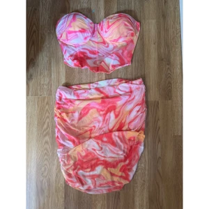 Färgglad tvådelad kortklänning - Tvådelad klänning med bandeau-topp och tight kjol i swirlmönster med rosa, orange och vit. Kjolen är kort och har rynkade detaljer. Perfekt för sommarens alla tillfällen när du vill sticka ut.