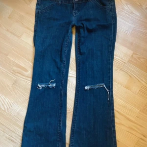 Mörkblå bootcut jeans med slitna knän - Säljer ett par mörkblå jeans med bootcut passform och slitna detaljer vid knäna. Jeansen har låg midja och klassisk femficksdesign. Perfekta för dig som gillar en avslappnad och trendig look. Materialet är jeans i bomull.