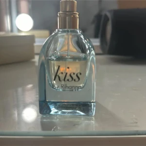 Kiss by Rihanna parfym - Fräsch parfym från Rihanna i en stilren, transparent glasflaska med ljusblå ton och guldig spraykork. Flaskan har en modern, fyrkantig form och texten 'Kiss by Rihanna' på framsidan. Perfekt för dig som gillar kändisdofter och snygg design.