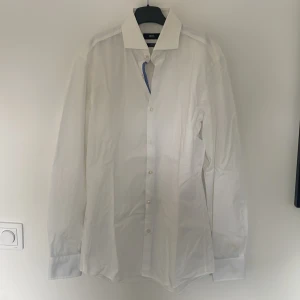Vit skjorta från BOSS st.37 - Klassisk vit skjorta från BOSS i slim fit-modell. Skjortan har långa ärmar, knappar framtill och en stilren krage. Perfekt för dig som gillar en clean och stilren look. Materialet är lätt att stryka och känns bekvämt mot huden.