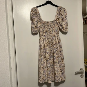 Blommig klänning med puffärm från H&M - Supersöt blommig klänning från H&M med smock upptill och puffiga kortärmar. Klänningen har en ljus bas med blå och beige blommor, och är tillverkad i mjuk polyester med lite stretch. Perfekt för vår och sommar!