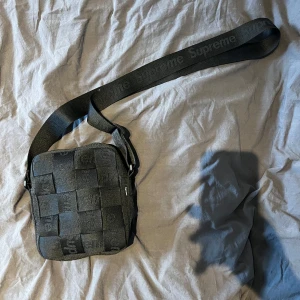 Svart Supreme axelväska - Supreme axelväska i svart med vävt rutmönster och justerbar axelrem där loggan är invävd. Väskan har en kompakt fyrkantig form och är perfekt för att bära det viktigaste. Snygg och stilren accessoar för streetwear. Väldigt bra skick