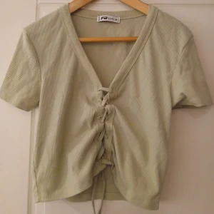 Ljusgrön ribbad topp med snörning från FB Sister - Säljer en ljusgrön ribbad topp från FB Sister i storlek M. Toppen har korta ärmar, v-ringning och snygg snörning framtill som ger en trendig look. Perfekt att matcha med jeans eller kjol!