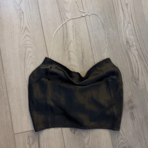 Svart fest topp med kedjedetalj från NA-KD - Säljer en svart linnetopp från NA-KD med snygg kedja som axelband och draperad framsida. Toppen har en croppad passform och är perfekt för att lyfta din outfit med en trendig touch till fest outfiten.