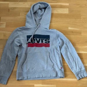  Levi's Hoodie - Säljer en klassisk grå hoodie från Levi's med stor logga i blått och rött framtill. Tröjan har huva med dragsko och en stor magficka. Perfekt för en avslappnad stil.