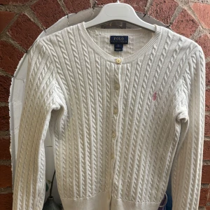 Vit kabelstickad kofta från Polo Ralph Lauren - Säljer en klassisk vit kabelstickad kofta från Polo Ralph Lauren med rund halsringning och knappar framtill. Tröjan har långa ärmar och en liten rosa broderad logga på bröstet. Perfekt att styla till olika outfits!