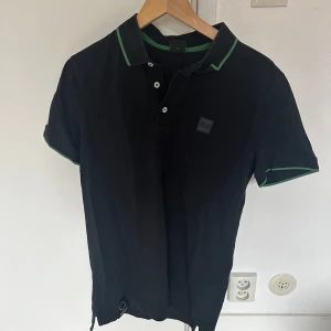 Svart pikétröja från Hugo Boss - Säljer en svart pikétröja från Hugo Boss med gröna detaljer på krage och ärmslut. Tröjan har korta ärmar, knappar framtill och klassisk pikékrage. Perfekt för en stilren och sportig look.