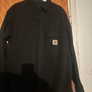 Carhartt WIP Reno Jacka - Säljer en svart skjorta från Carhartt med klassisk krage och knappstängning framtill. Skjortan har en bröstficka med Carhartt-logga och långa ärmar. Perfekt för dig som gillar stilrena och enkla plagg. Nypris 1600kr (Zalando)