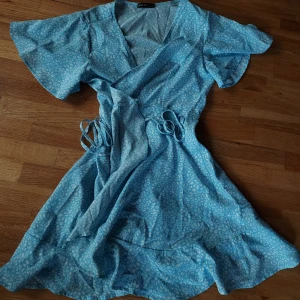 Ljusblå omlottklänning med mönster - Säljer en ljusblå omlottklänning med vitt småmönster. Klänningen har korta, vida ärmar och knyt i midjan för en fin passform. Perfekt för varma dagar och har en lätt, luftig känsla. Klänningen har en ganska märklig knytning men är super lätt när man fattat den. Skriv priv för förklaring på hur man knyter den:)