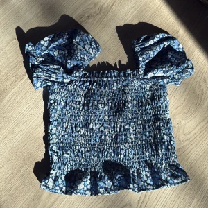 Blommig smockad topp med puffärmar från Uniq Young - Supersöt blå smockad topp med vita blommor och volangkant. Toppen har korta puffärmar och är stretchig över hela kroppen. Perfekt för en somrig look!