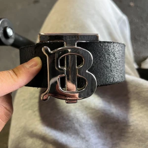 Svart skärp med silverfärgat TP märke - Säljer ett svart skärp i skinn med ett stort, silverfärgat TB-spänne från Burberry. Bältet är brett och har en stilren, lyxig känsla med tydlig logga framtill. Perfekt accessoar för att lyfta din outfit.