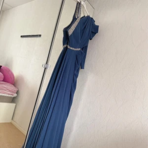 Blå långklänning med strass från Bellisse - Säljer en blå långklänning från Bellisse med enaxlad design och glittriga strassdetaljer vid axel och midja. Klänningen har ett elegant fall och lång ärm på ena sidan, perfekt för speciella tillfällen. Använd endas 1 gång