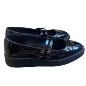 Svarta lackade Mary Jane ballerinaskor - Säljer ett par svarta glansiga Mary Jane ballerinaskor med dubbla remmar och spännen. Skorna har rund tå och platt sula, perfekt för dig som gillar klassisk och stilren look.