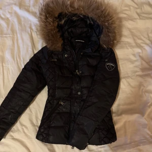 Svart dunjacka med fuskpäls från Rockandblue - Säljer en svart dunjacka från Rockandblue med avtagbar huva och fuskpäls. Jackan har quiltade partier, dragkedjor och en snygg badge på ärmen. Perfekt för kyliga dagar och har en figurnära passform.