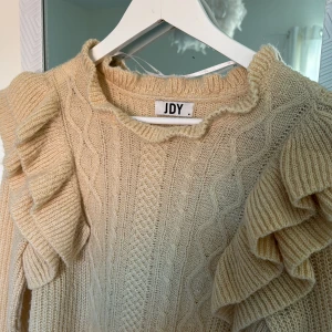 Beige stickad tröja med volang från JDY - Säljer en beige stickad tröja från JDY med volangdetaljer längs axlarna och kabelstickat mönster framtill. Tröjan har långa ärmar och en rund halsringning. Skriv om ni har några frågor!!💕