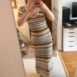 Stickad långklänning med mönster - Säljer en stickad långklänning med kort ärm och v-ringning. Klänningen har ett färgglatt randigt mönster i beige, vit, brun, blå och gul. Den är figurnära och har virkade detaljer vid halsringningen. 