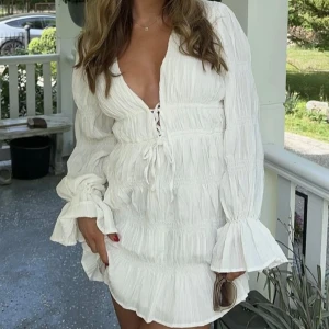 Vit volangklänning med v-ringning och knyt NAKD - Superfin vit klänning med djup v-ringning, långa volangärmar och knyt framtill. Moa Mattsson x  NAKD