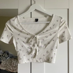 Vit blommig topp - Säljer en söt vit croppad topp med små lila blommor och gröna blad. Toppen har korta ärmar, ribbad struktur, v-ringning och knytning framtill. Perfekt för varma dagar!🩷🥰
