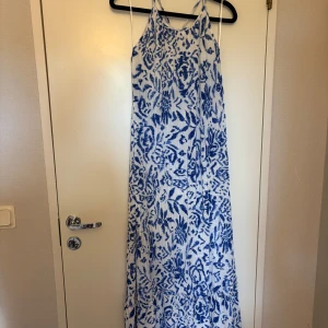 Vit och blå långklänning med mönster - Säljer en somrig långklänning i vitt med blått mönster. Klänningen har smala axelband och en rak, luftig passform. Perfekt för varma dagar och semester. Mönstret är inspirerat av blommor och blad.
