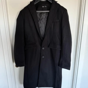 Svart rock - Stilren svart rock från Manfinity med klassisk krage och två knappar framtill. Jackan har långa ärmar och två fickor på sidorna samt en innerficka. Perfekt för kyligare dagar.