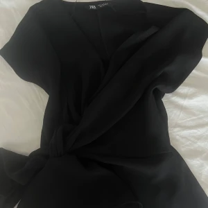 Svart omlottblus från Zara - Snygg svart omlottblus från Zara med korta ärmar och knyt i midjan. Perfekt för en stilren look och enkel att matcha med olika underdelar.
