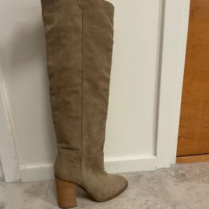 Beiga höga stövlar med klack - Säljer ett par snygga höga stövlar i beige mocka med rund tå och kraftig klack. Stövlarna går över knäet och har en stilren design som passar perfekt till många outfits.