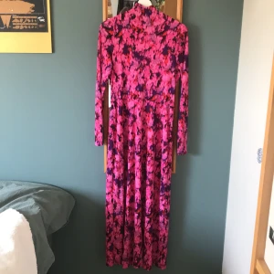 Långklänning med blommigt mönster i rosa och lila - Säljer en långklänning med blommigt mönster i rosa och lila toner. Klänningen har lång ärm och hög hals, perfekt för dig som vill sticka ut med färg. Materialet känns mjukt och följsamt.