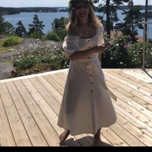 Vit klänning med puffärm och knappar - Säljer en söt vit klänning med puffärmar och knappar framtill. Klänningen har knytband i midjan och är tillverkad i ett lätt bomullstyg. Perfekt för varma dagar och har en feminin siluett med fina detaljer. Perfekt till midsommar eller en sommarkväll, stolen xs använd på midsommar en gång!