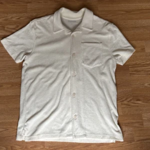 Vit frottéskjorta från Kappahl - Säljer en vit kortärmad skjorta från Kappahl i mjuk frotté. Skjortan har klassisk krage, bröstficka och knäppning framtill. Perfekt för en avslappnad och stilren look. Nypris 500kr