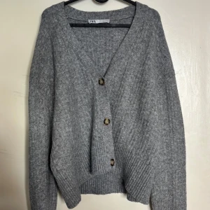 Grå stickad kofta från Zara - Mysig grå stickad kofta från Zara med v-ringning och tre stora knappar framtill. Koftan har lång ärm och ribbade detaljer vid ärmslut och nederkant. Perfekt för lager-på-lager under kyliga dagar.                                Använd fåtal gånger