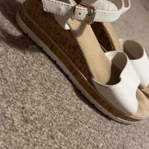Vita sandaler från Graceland med korksula - Säljer ett par vita sandaler från Graceland med bred rem över tårna och justerbart spänne vid ankeln. Skorna har en bekväm korksula och platt vit yttersula. Perfekta för varma dagar och enkel att matcha med sommarens outfits.