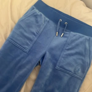 Blå mjukisbyxor från Juicy Couture - Köpte den för 1000kr och har bara använt den 1 gång. Säljer ett par blå mjukisbyxor från Juicy Couture med bred resår i midjan och dragsko med metalländar. Byxorna har stora fickor fram och bak, samt broderad JC-logga på ena fickan. Mjukt och glansigt material som känns lyxigt.
