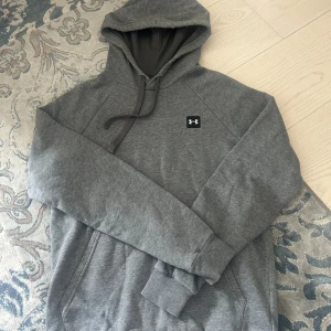 Grå hoodie från Under Armour - Säljer en grå hoodie från Under Armour med klassisk huva och dragsnören. Tröjan har en liten logotyp på bröstet och en stor magficka framtill. Perfekt för en avslappnad stil.