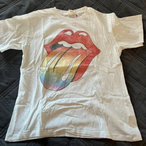 Vit Rolling Stones t-shirt från H&M - Vit t-shirt från H&M med det klassiska Rolling Stones-trycket framtill i rött och regnbågsfärger. Baksidan har texten 'The Rolling Stones' i svart. T-shirten har rund hals och korta ärmar. Perfekt för dig som gillar bandmerch och vill ha en cool look.