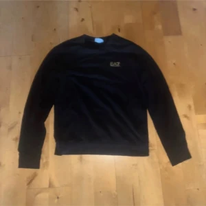 Svart Ea7 tröja - Säljer en svart sweatshirt från Emporio Armani EA7 med guldfärgad logga på bröstet. Tröjan har rund halsringning och långa ärmar. Använd fåtal gånger. Säljs eftersom jag har bytt stil. Pris kan diskuteras  
